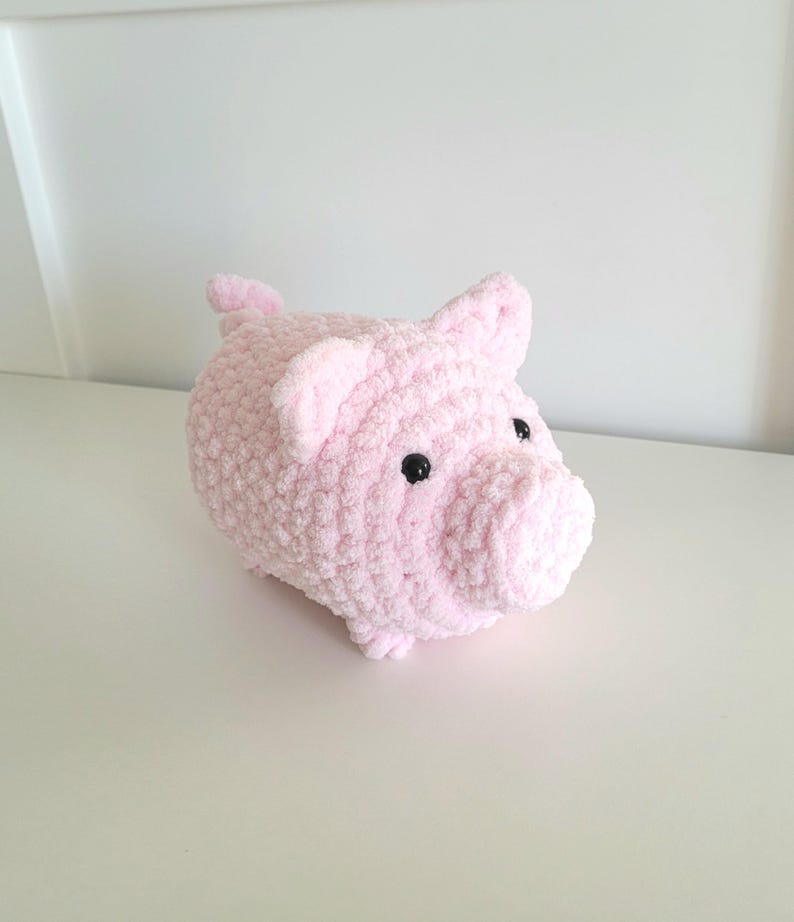 May include: A plush, light pink pig with black button eyes and small ears. The handmade piggy bank is made of soft, textured yarn. The pig is sitting on a white surface.
