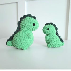 Puede incluir: Dos peluches de dinosaurio verde hechos a crochet con ojos negros. El dinosaurio más grande está de pie, mientras que el más pequeño está sentado.