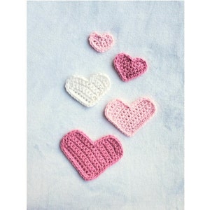 Puede incluir: Cinco corazones tejidos a crochet en varios tonos de rosa y blanco. Los corazones están dispuestos en forma de pirámide, con el corazón más grande en la parte inferior y el corazón más pequeño en la parte superior.