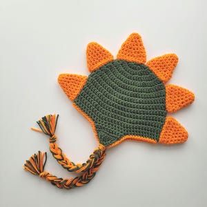 Puede incluir: Un gorro de dinosaurio de crochet verde y naranja con una larga cola trenzada.