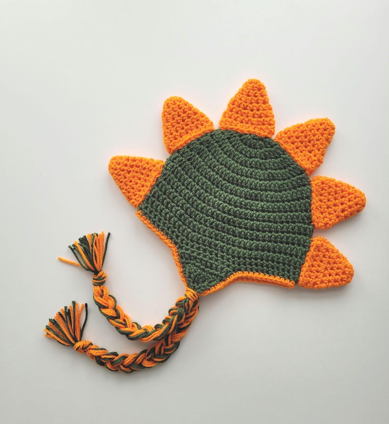 Puede incluir: Un gorro de dinosaurio de crochet verde y naranja con una larga cola trenzada.