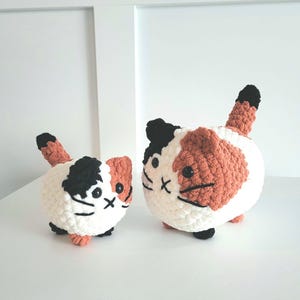 May include: Two crocheted cat plushies, one larger than the other. Both are white with black and orange patches, and have black eyes and whiskers.