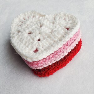 May include: A crocheted heart-shaped coaster in white, pink, and red. The coaster is made of multiple layers of yarn, creating a layered effect.
