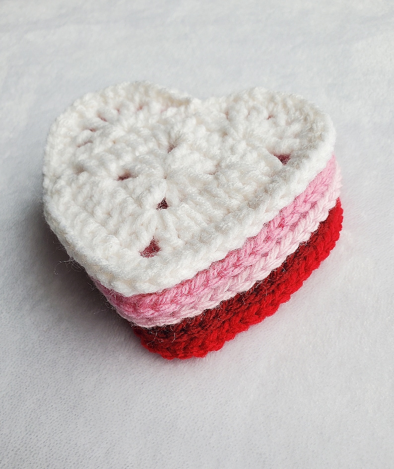May include: A crocheted heart-shaped coaster in white, pink, and red. The coaster is made of multiple layers of yarn, creating a layered effect.