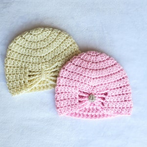 Puede incluir: Dos gorros de ganchillo, uno en amarillo pálido y otro en rosa brillante, ambos con un lazo decorativo y un acento de joya.