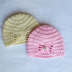 May include: Two crocheted hats, one in a pale yellow and one in a bright pink. Both hats have a bow detail and a small jeweled embellishment.