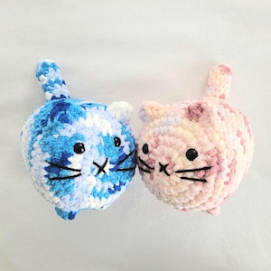 May include: Two crocheted cat plushies, one blue and white and one pink and white, facing each other.