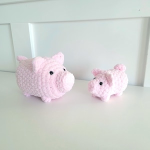 May include: Two handmade pink crochet pig figurines with black eyes. The larger pig has a rounded body, ears, and snout. The smaller pig has a similar design. Both are sitting on a white surface.