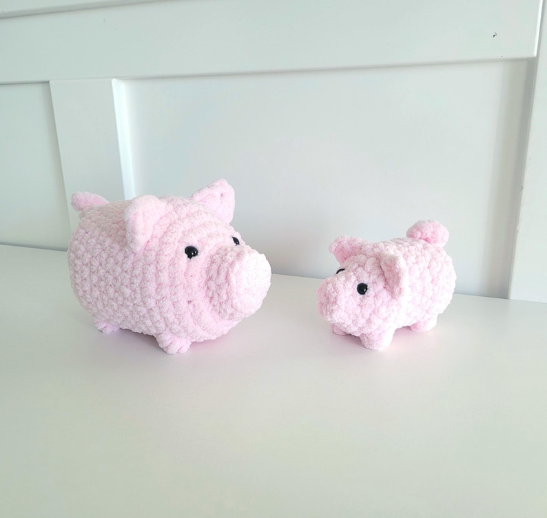 May include: Two handmade pink crochet pig figurines with black eyes. The larger pig has a rounded body, ears, and snout. The smaller pig has a similar design. Both are sitting on a white surface.