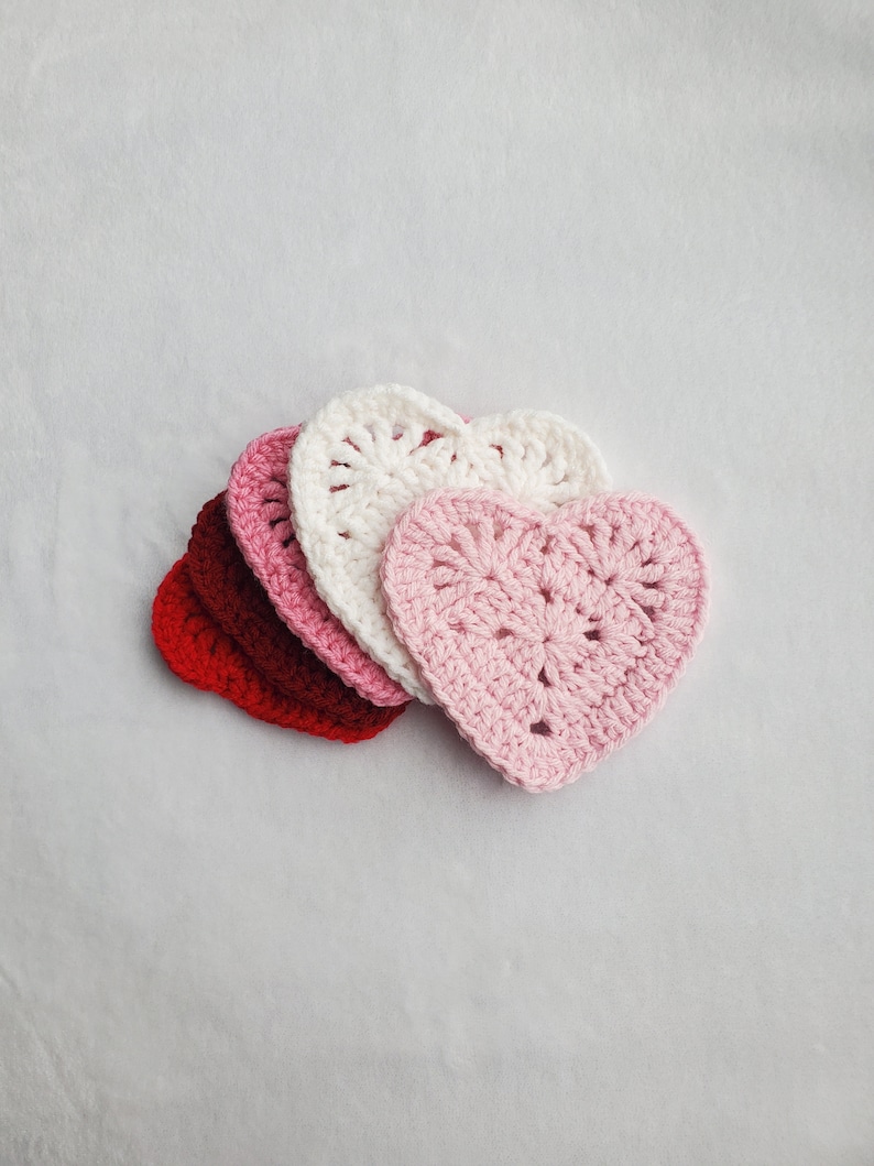 May include: Four crocheted heart-shaped coasters in shades of red, pink, and white. The coasters are stacked on top of each other.