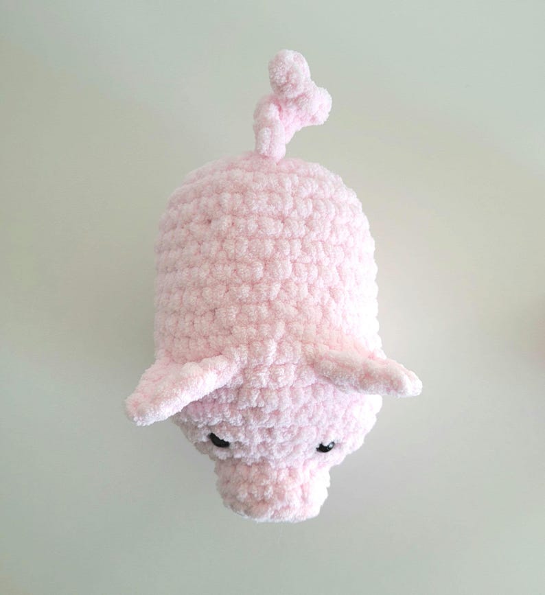 May include: A handmade, light pink crocheted pig with small black eyes, floppy ears, and a curly tail. The plush toy is made of soft, textured yarn, and is a whimsical, cuddly creation.