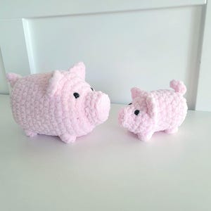 May include: Two pink crochet pig figurines. The larger pig has a rounded body, small ears, and black eyes. The smaller pig has a similar design. Both are sitting on a white surface.
