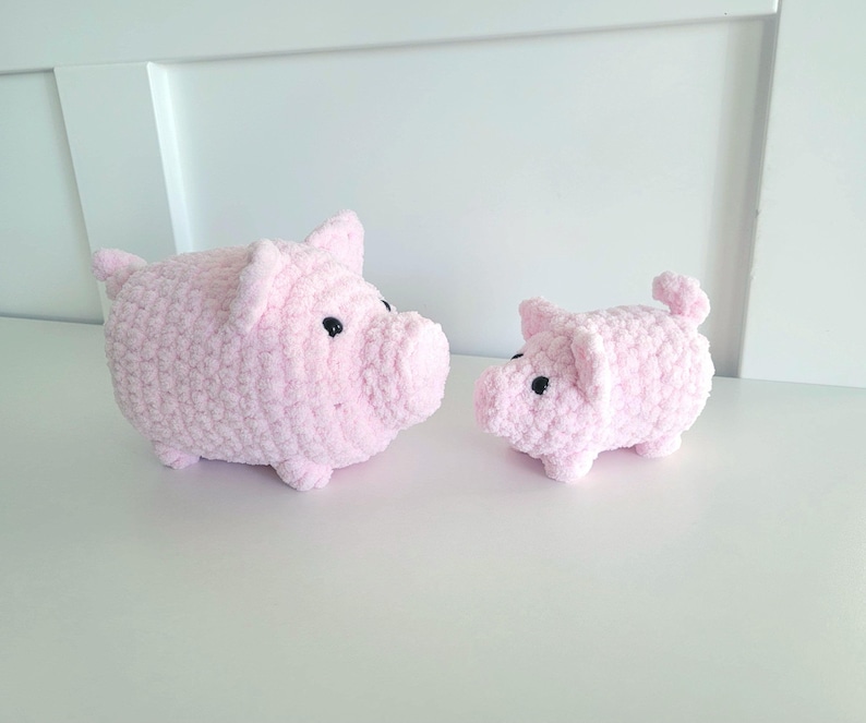 May include: Two pink crochet pig figurines. The larger pig has a rounded body, small ears, and black eyes. The smaller pig has a similar design. Both are sitting on a white surface.