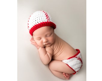 May include: A white and red crocheted hat and matching diaper cover set. The hat features a brim and a baseball-inspired pattern with red stitching details. The diaper cover is white with red trim and matching red stitching accents.