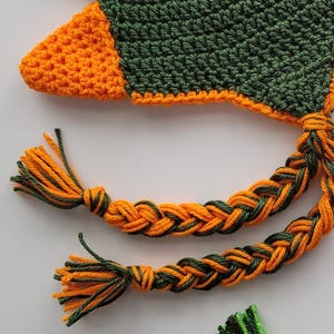 Puede incluir: Un gorro de crochet con un borla trenzada verde y naranja. El gorro est&aacute; hecho de un hilo suave y texturizado y tiene un dise&ntilde;o &uacute;nico.