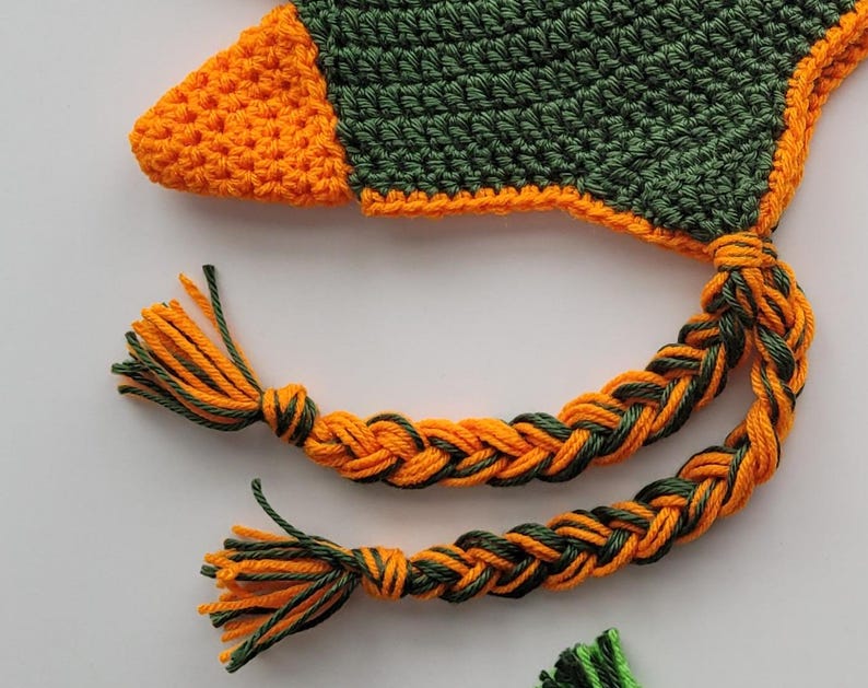 Puede incluir: Un gorro de crochet con un borla trenzada verde y naranja. El gorro est&aacute; hecho de un hilo suave y texturizado y tiene un dise&ntilde;o &uacute;nico.