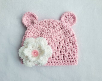 Op de afbeelding: Roze gebreide baby muts met berenoren en een witte gehaakte bloem met een roze knoop in het midden.