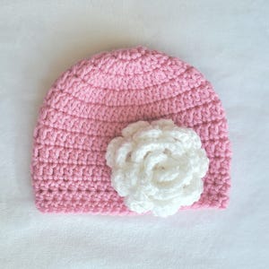 Pode incluir: Um gorro de crochê rosa com uma flor de crochê branca no topo.