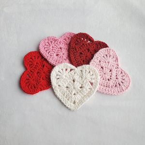 May include: Five crocheted hearts in different colours: red, pink, white. The hearts are arranged in a semi-circle on a white background.