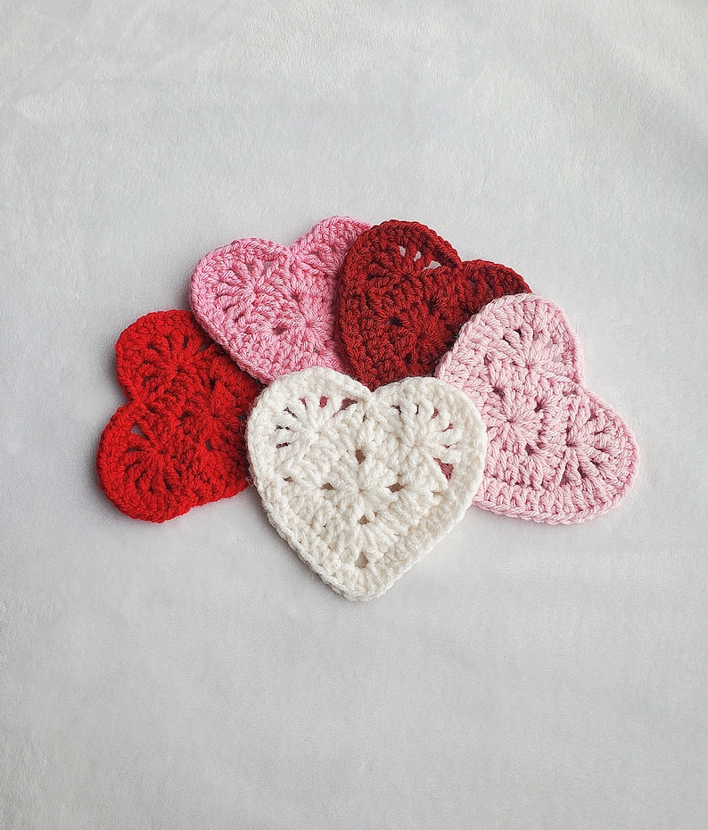 May include: Five crocheted hearts in different colours: red, pink, white. The hearts are arranged in a semi-circle on a white background.