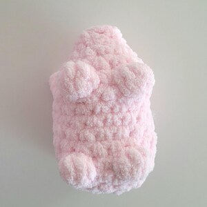 May include: A light pink, plush, handmade stuffed animal in the shape of a bear. The bear has a textured, fuzzy appearance with rounded paws and a body. The toy is made of a soft, fluffy material.