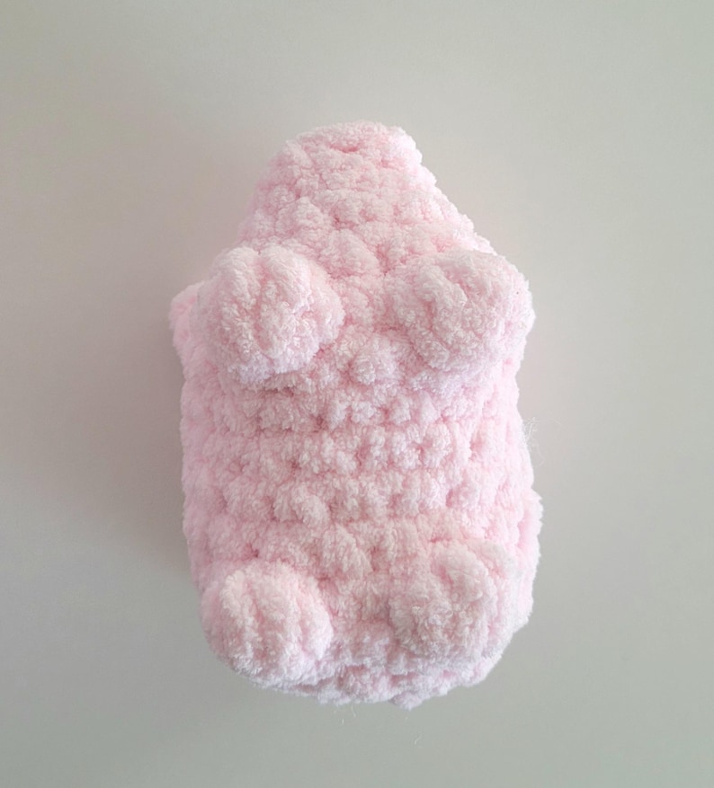 May include: A light pink, plush, handmade stuffed animal in the shape of a bear. The bear has a textured, fuzzy appearance with rounded paws and a body. The toy is made of a soft, fluffy material.