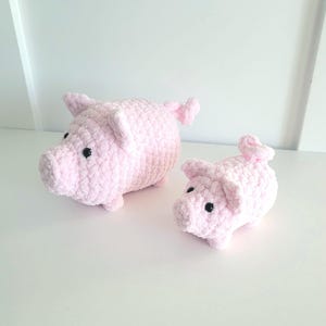 May include: Two handmade pink crochet pig figurines. The larger pig has a rounded body, small ears, and a curly tail. The smaller pig has similar features. Both have black button eyes and are made of soft, textured yarn.