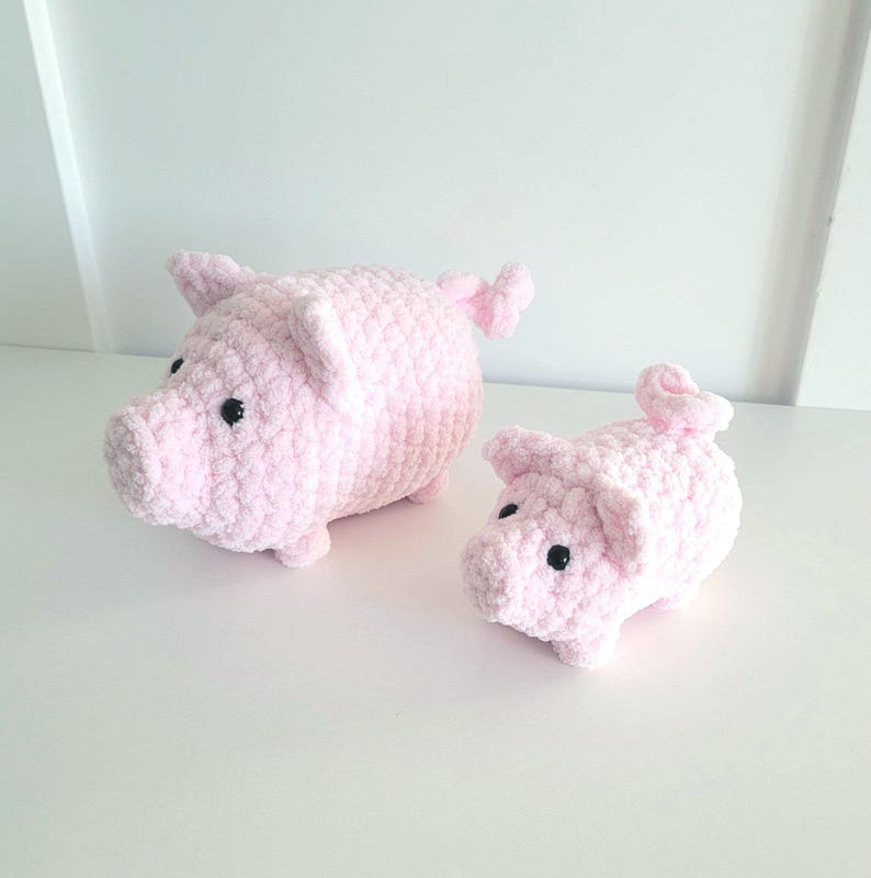 May include: Two handmade pink crochet pig figurines. The larger pig has a rounded body, small ears, and a curly tail. The smaller pig has similar features. Both have black button eyes and are made of soft, textured yarn.