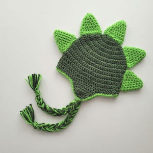Puede incluir: Sombrero de dinosaurio de crochet verde y verde oscuro con una larga cola trenzada.