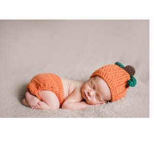 May include: A newborn baby wearing an orange crocheted pumpkin hat and matching diaper. The baby is sleeping on a light brown fuzzy blanket.