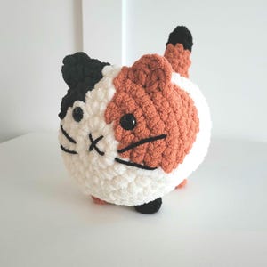 Op de afbeelding: Een gehaakt knuffeldier in de vorm van een kat met zwart, wit en oranje bont. De kat heeft zwarte ogen en een zwarte neus.