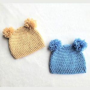 May include: Two crocheted hats with pom-poms. One hat is beige and the other is light blue.
