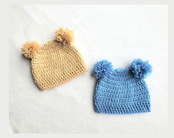 Puede incluir: Dos gorros de punto con pompones. Un gorro es beige y el otro es azul claro.