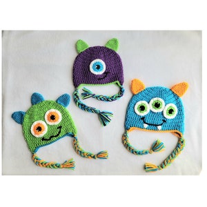 May include: Three crocheted monster hats with different colored fur and faces. The hats have long, braided ties. One hat is purple with a single eye, one is green with two eyes, and one is blue with three eyes.
