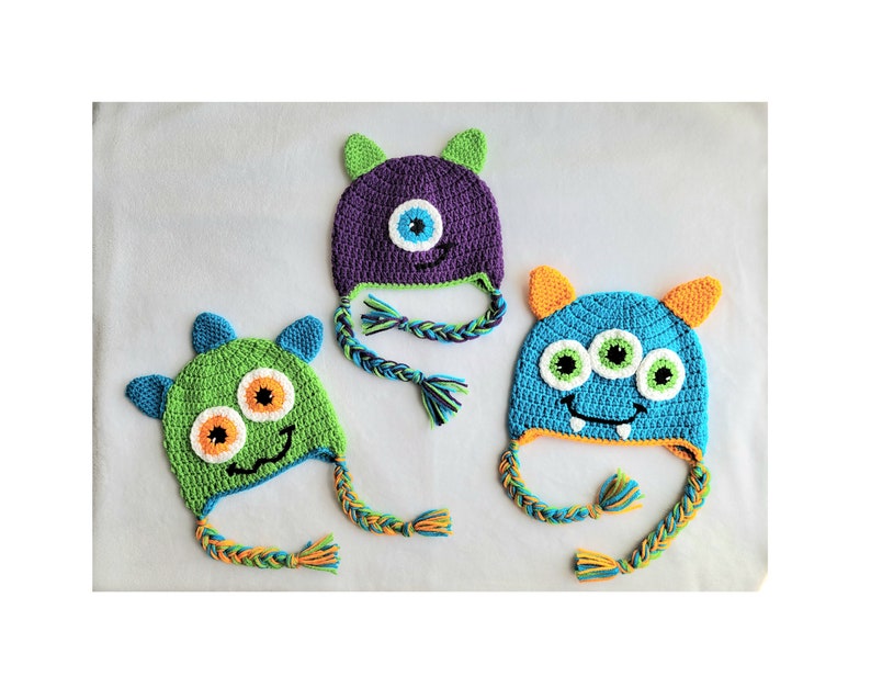May include: Three crocheted monster hats with different colored fur and faces. The hats have long, braided ties. One hat is purple with a single eye, one is green with two eyes, and one is blue with three eyes.