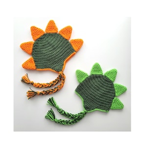 May include: Two crocheted dinosaur hats with green and orange spikes. The hats have long braided ties.