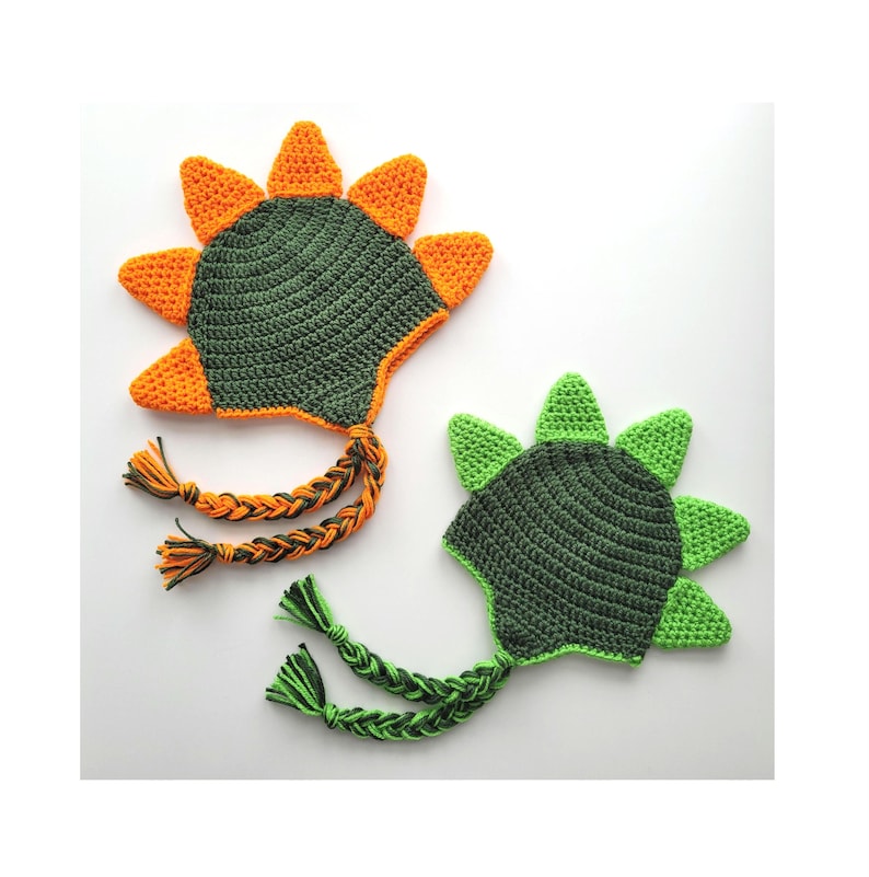 Puede incluir: Dos gorros de dinosaurio de crochet con p&uacute;as verdes y naranjas. Los gorros tienen largas ataduras trenzadas.