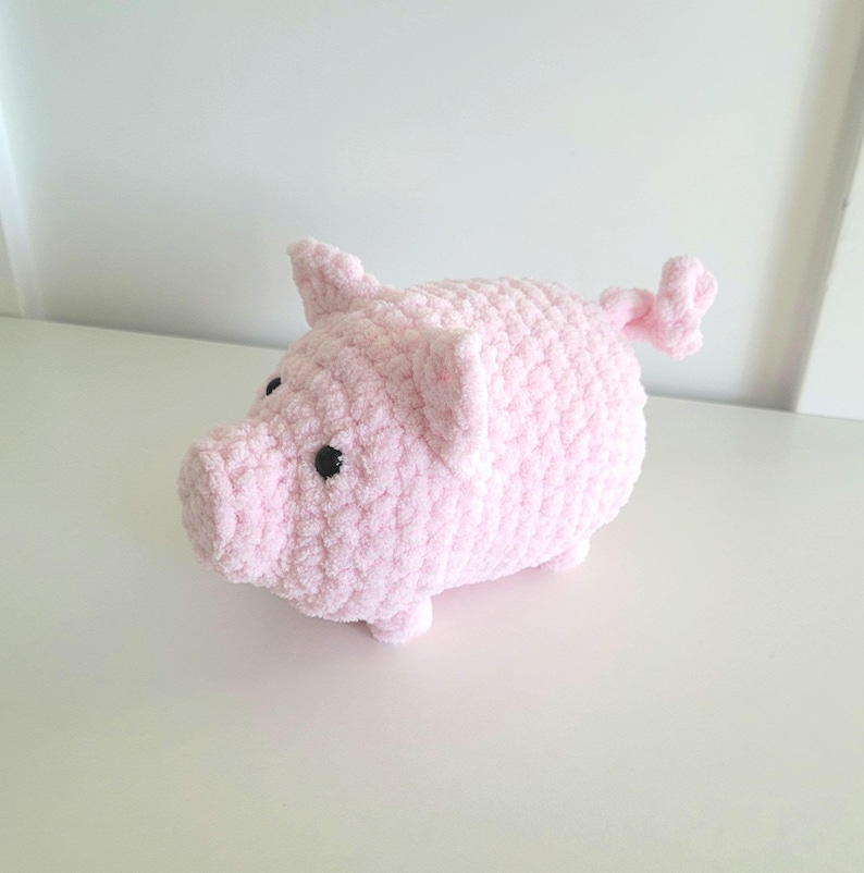 May include: A handmade, plush pink pig with black button eyes and a curly tail. The pig has small ears and four tiny legs. This soft, cuddly toy is perfect for a nursery or as a decorative item.