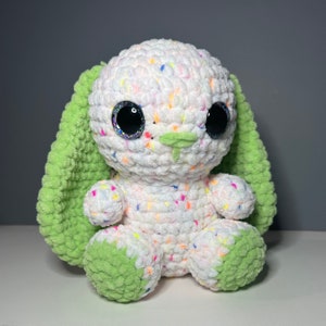 Może przedstawiać: Ręcznie robiony, pluszowy króliczek z białym korpusem nakrapianym kolorowymi plamkami. Króliczek ma duże, błyszczące czarne oczy, zielone wewnętrzne uszy i zielone akcenty na łapkach. Miękka zabawka dla dzieci.