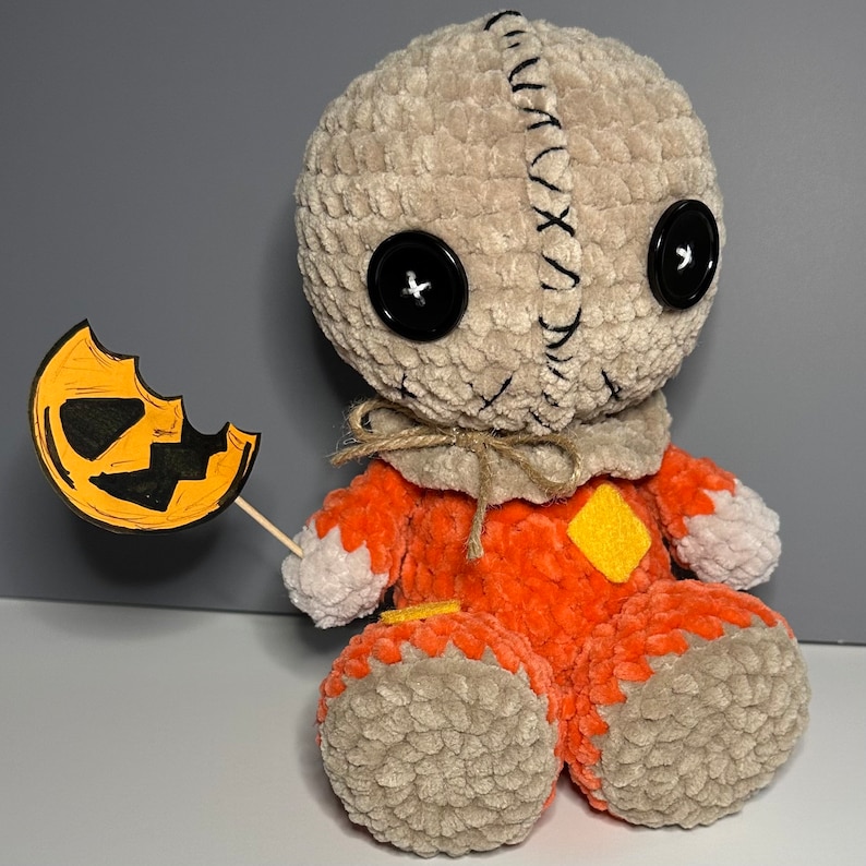 Puede incluir: Un juguete de peluche tejido a crochet que se asemeja a un personaje de la pel&iacute;cula "Pesadilla antes de Navidad". El juguete lleva un disfraz marr&oacute;n y naranja con una cara cosida y ojos de bot&oacute;n negros. El juguete sostiene un peque&ntilde;o recorte de calabaza naranja y negro.