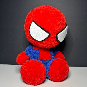 Peut inclure: Un jouet en peluche crochet&eacute; rouge et bleu repr&eacute;sentant un super-h&eacute;ros avec des yeux blancs. Le jouet est assis sur une surface grise.