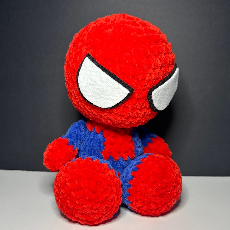 Peut inclure: Un jouet en peluche crochet&eacute; rouge et bleu repr&eacute;sentant un super-h&eacute;ros avec des yeux blancs. Le jouet est assis sur une surface grise.