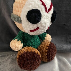 Puede incluir: Un juguete de peluche tejido a crochet con una m&aacute;scara blanca, marcas rojas y ojos negros. El juguete lleva una camisa verde y tiene piernas marrones.