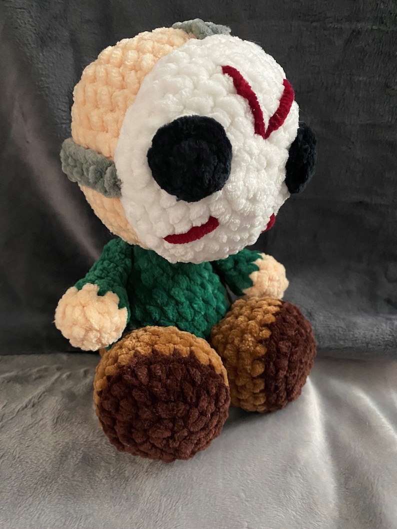 Puede incluir: Un juguete de peluche tejido a crochet con una m&aacute;scara blanca, marcas rojas y ojos negros. El juguete lleva una camisa verde y tiene piernas marrones.