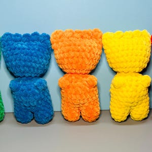 May include: Five crocheted teddy bears in a row, from left to right: green, blue, orange, yellow, and red.