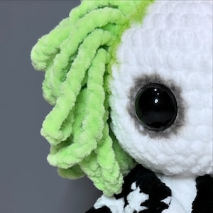 May include: Close-up of a crocheted doll with green yarn hair and a large black eye. The doll is wearing a black and white striped shirt.