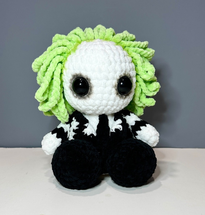 May include: A crocheted plush toy with a white face, black eyes, and green hair. The toy is wearing a black and white striped jacket and black pants.