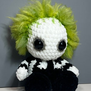 May include: A crocheted plush toy resembling a character with green hair, black eyes, and a black and white striped shirt. The toy is sitting with its legs crossed.