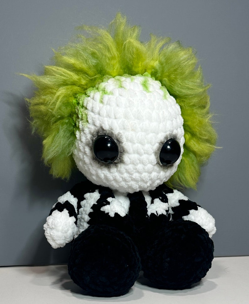 May include: A crocheted plush toy resembling a character with green hair, black eyes, and a black and white striped shirt. The toy is sitting with its legs crossed.