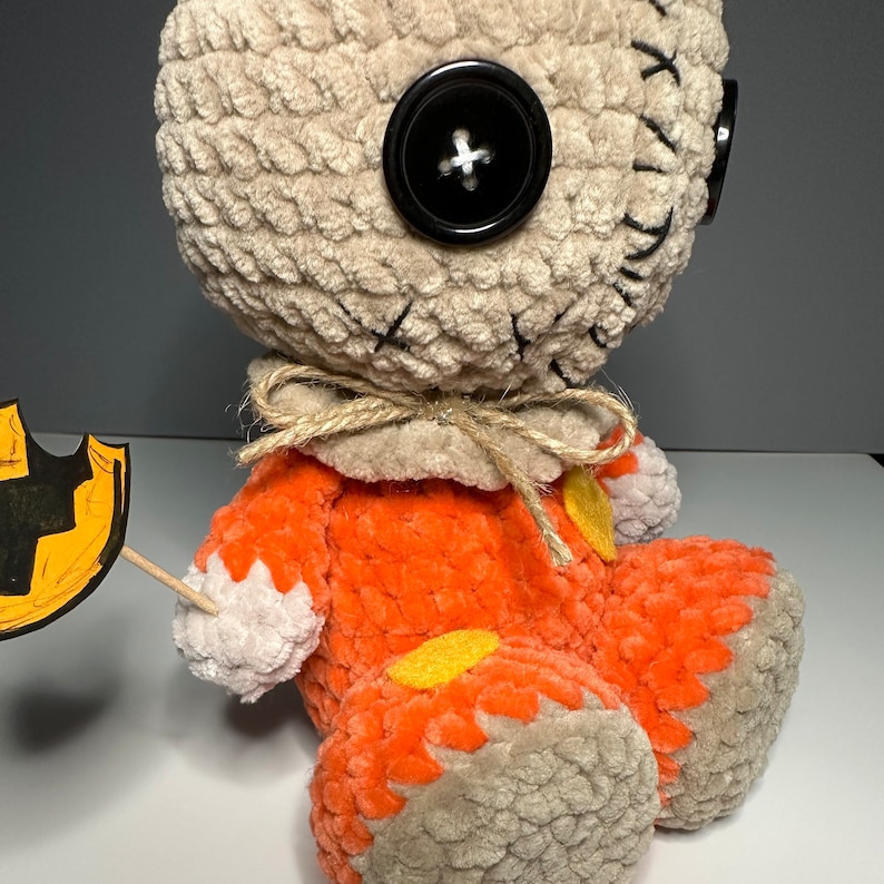 Puede incluir: Un juguete de peluche tejido a crochet con forma de monstruo cosido que lleva un mono naranja con detalles amarillos. El juguete tiene un bot&oacute;n negro grande como ojo y sostiene un peque&ntilde;o cartel naranja y negro con tem&aacute;tica de Halloween.
